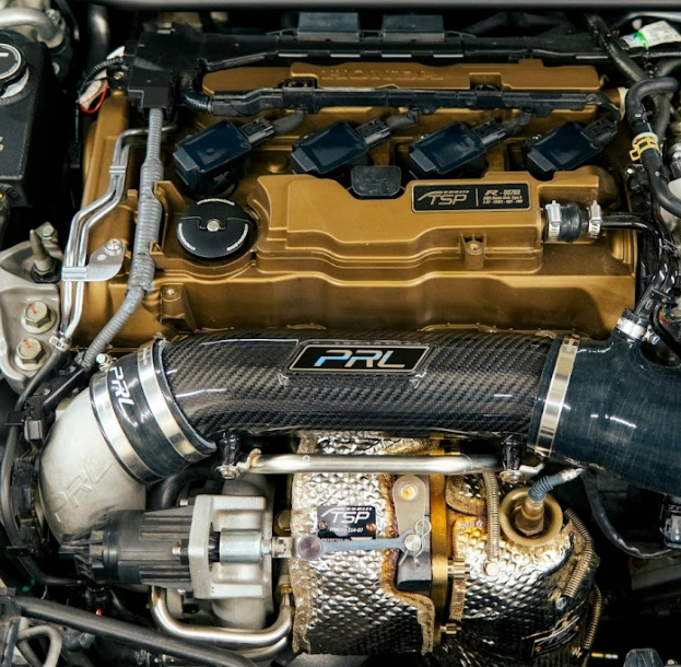 A turbocharger inlet pipe kit installed on the engine of a vehicle, featuring a carbon fiber pipe and silicone couplers.